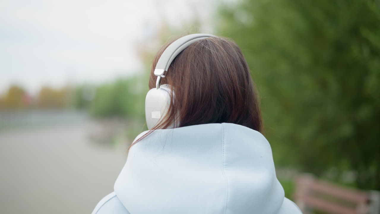 Back view of young woman wearing headphones, listening to music while strolling in garden with blurred trees in background. Ideal for videos depicting relaxation, music, and nature walks