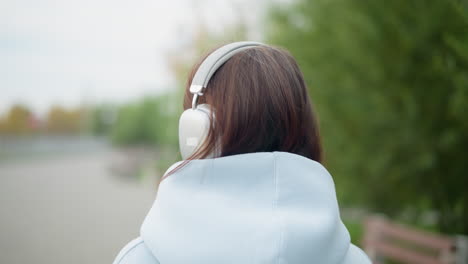 back view of young woman wearing headphones, listening to music while strolling in garden with blurred trees in background. ideal for videos depicting relaxation, music, and nature walks