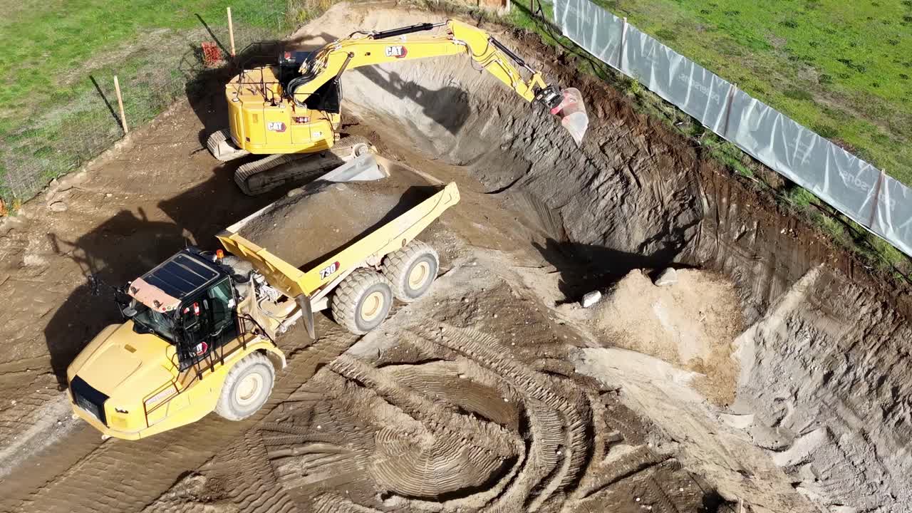 Aerial view of an excavator transferring soil into a dump truck at a construction site with green surroundings.
