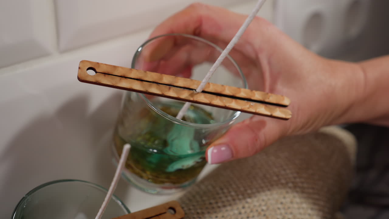Close-up of hand holding glass cup with greenish liquid and wick, dropping it near another glass cup in candle-making process, wooden sticks balance the wick as liquid is poured into the cup
