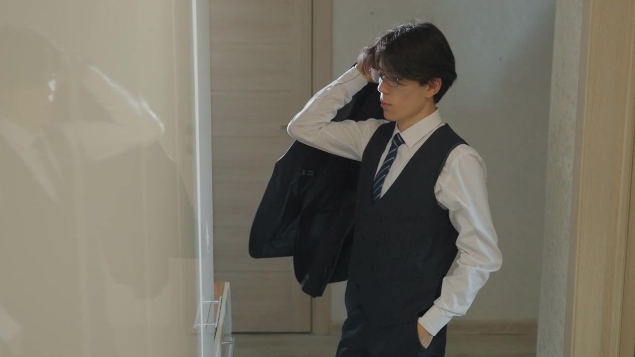 廊下でスーツを整える若い男性。鏡に映る自分を見ながらネクタイとベストを直し、ジャケットのボタンを留め、携帯電話を確認する。柔らかな朝の光がドアと廊下に温かい色合いを落とす。
