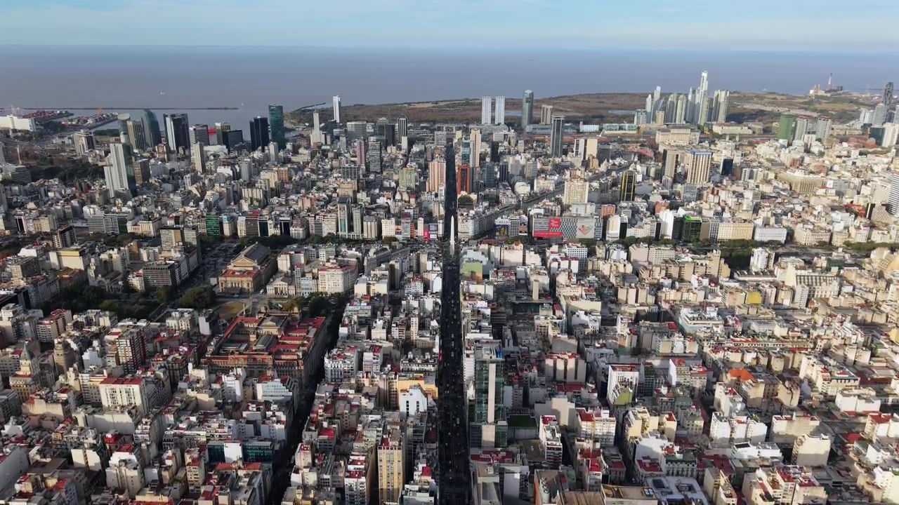 Ripresa aerea con drone in discesa sopra l'Obelisco e l'Avenida Corrientes a Buenos Aires, Argentina, che rivela la fitta griglia urbana, le torri finanziarie e il lungofiume del Río de la Plata.