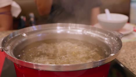 Opening-a-hotpot-filled-with-bubbling-boiling-broth-stock-for-a-mixed-vegetable-and-meat-soup,-inside-a-restaurant-in-Bangkok,-Thailand