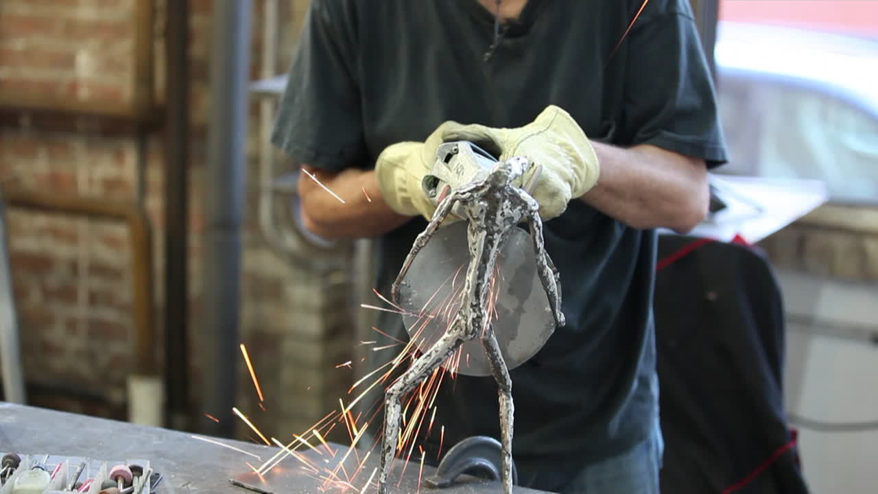 A man wearing gloves works on a small metal armature with a rotary grinder, sending showers of sparks toward the camera.