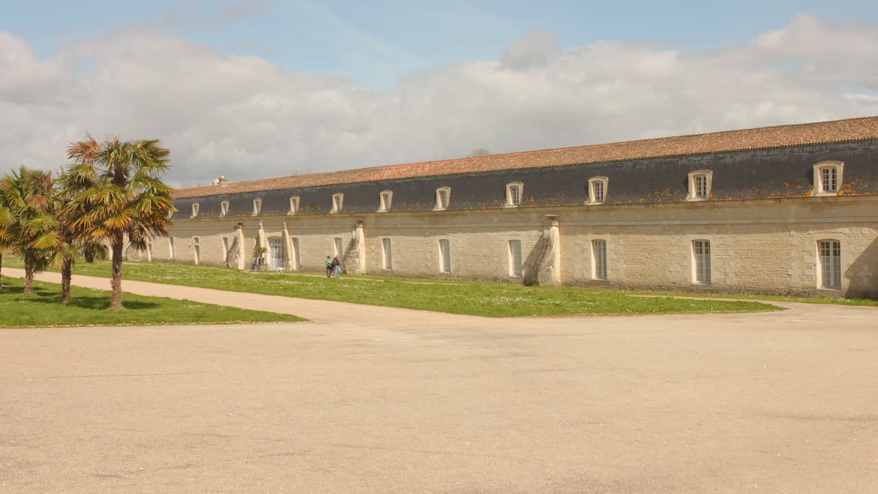 Historic stone building and palm trees in Rochefort's Corderie Garden under a bright sky