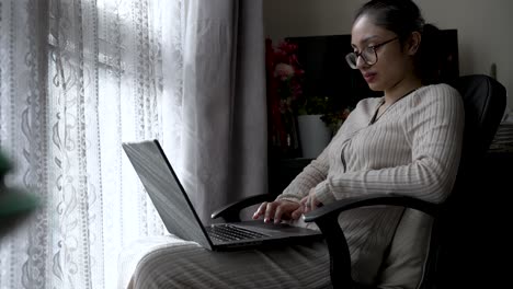 Asian-British-Housewife-Sitting-Near-Window-typing-on-laptop-placed-on-their-lap,-showcasing-the-integration-of-digital-media-into-home-environments