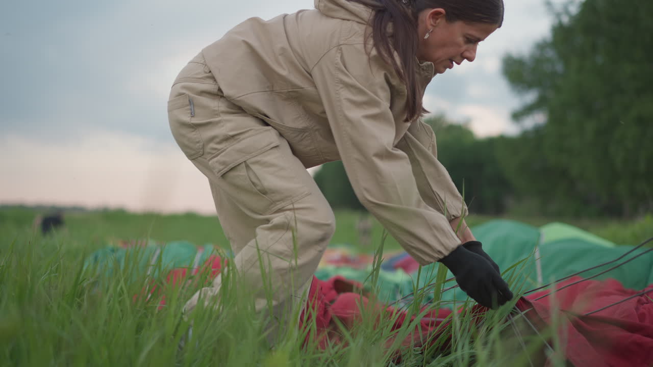 woman in flight suit crouching in grassy meadow handling hot air balloon envelope fabric and suspension cables during preflight setup at sunrise with forest backdrop and gentle breeze