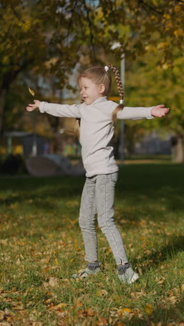 playful preschooler girl with long braids spins scattering yellow leaves. active child dances standing on green grass in park. happy childhood