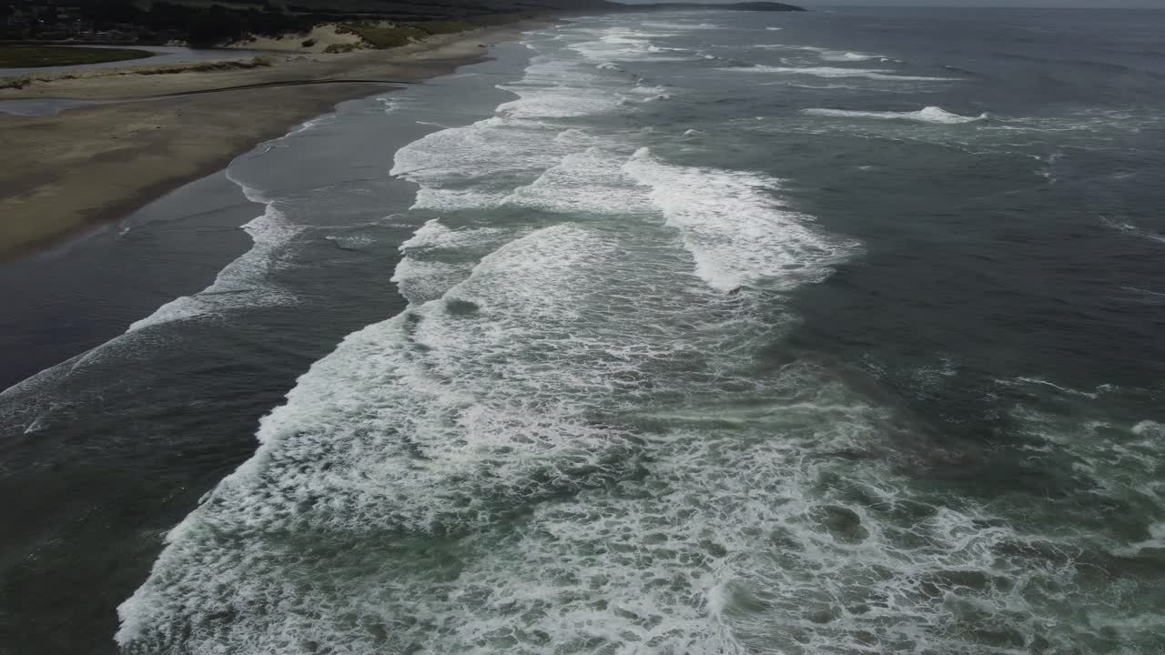 Aerial zoom shot of white waves crashing onto a sandy beach off the northern coast of California. Filmed on a cloudy summer day with a 4k drone.