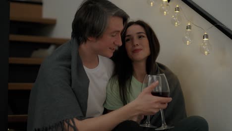 Portrait-of-a-couple-sit-on-the-wooden-stairs-hold-glasses-drink-red-wine-talk-laugh-hugging