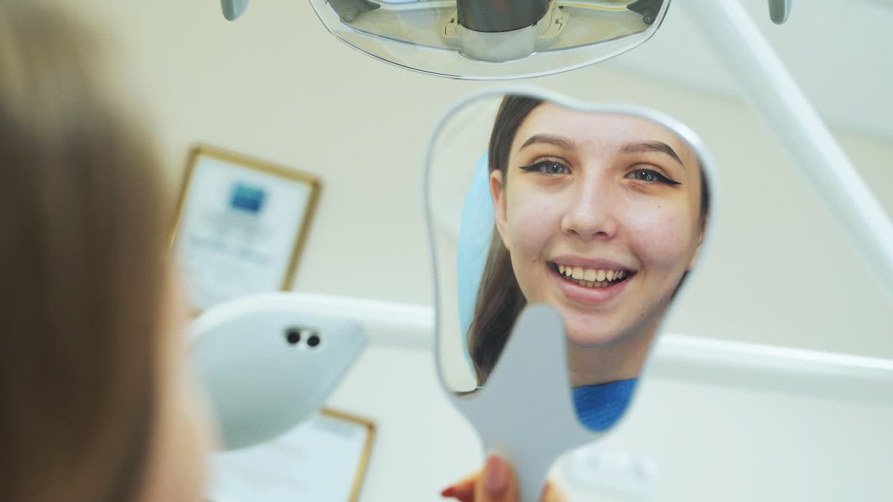 A young girl looks in the mirror in her hands as a dentist treats teeth in a clinic
