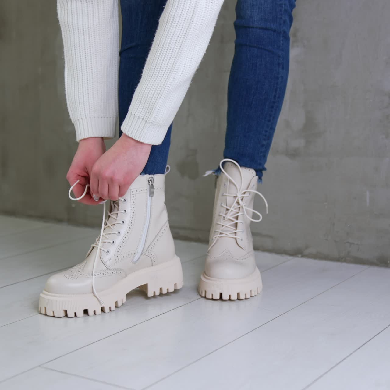 Unrecognized lady wearing jeans and white sweater bent to tie her lace on white boots. Model stands near the grey wall