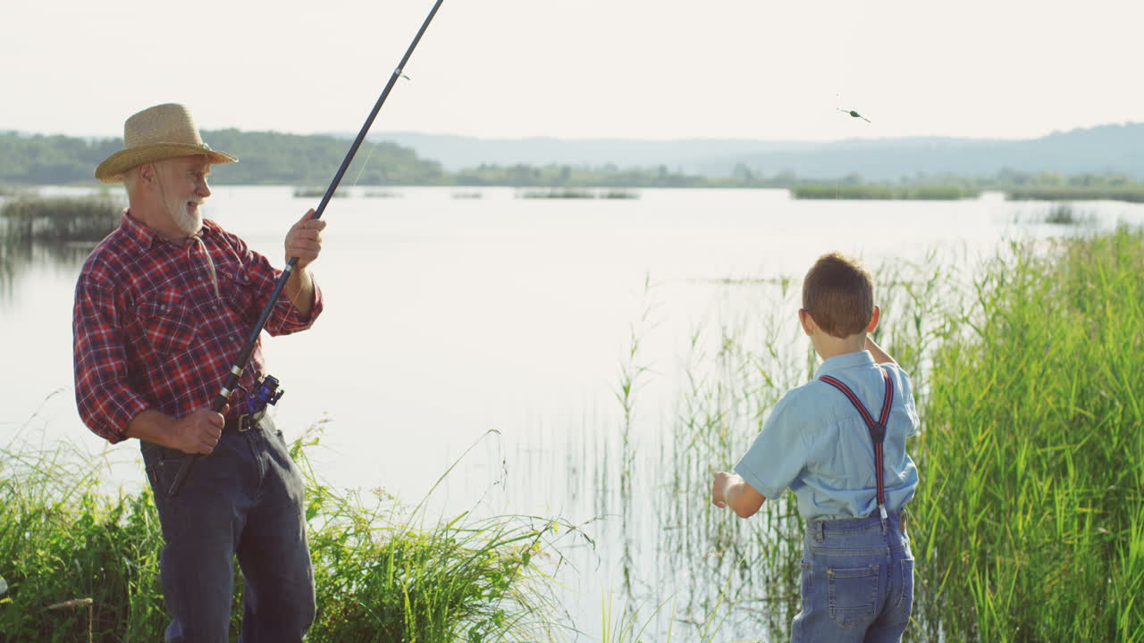 낚싯대를 들고 호수에서 물고기를 잡는 모자를 쓴 백인 노인과 그의 어린 아들이 물고기를 갈고리에서 떼고 있다