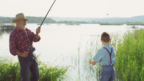 senior caucasian man in a hat catching a fish from the lake with a rod and his little son taking the fish off the hook