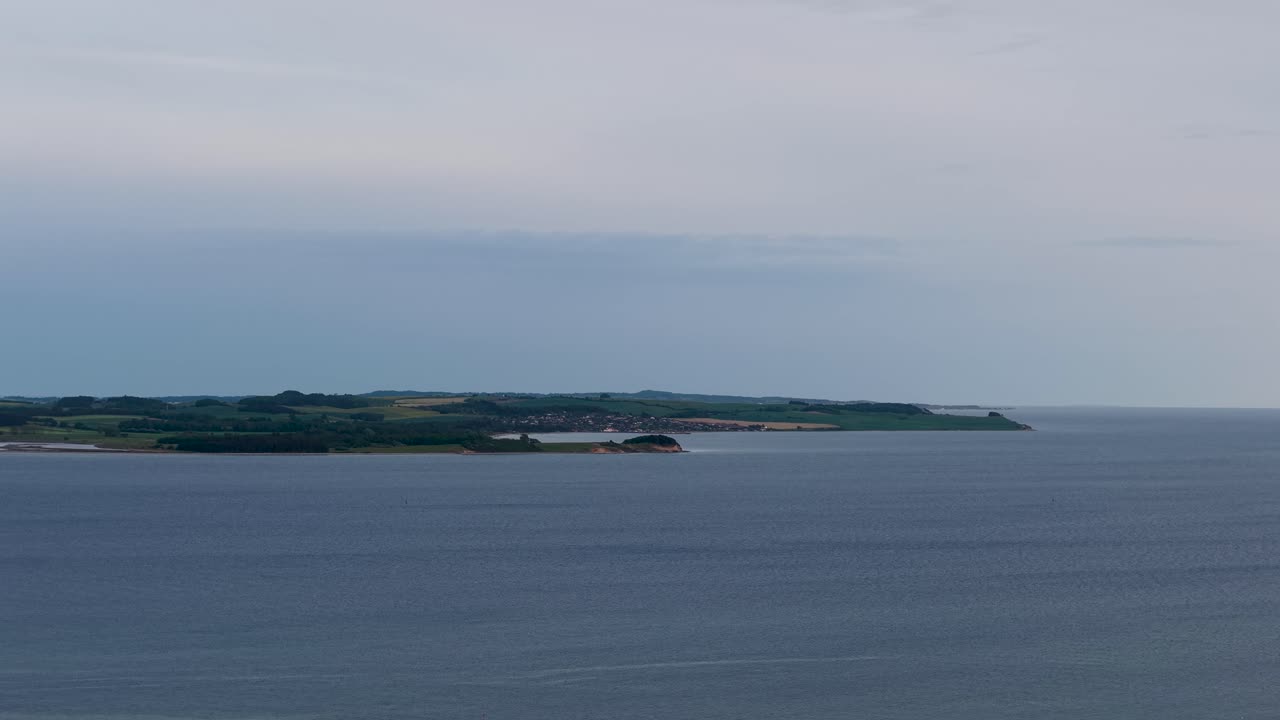 Drone footage captures a narrow island or peninsula along a calm blue sea in Denmark. The long landmass with green fields sits against the horizon under a cloudy sky