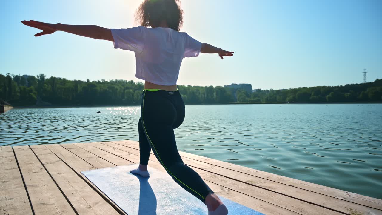 Young curly woman practicing yoga on the mat while wearing black protective mask. Park and lake in background. Sunny day. Corona Virus idea. Chisinau, Moldova