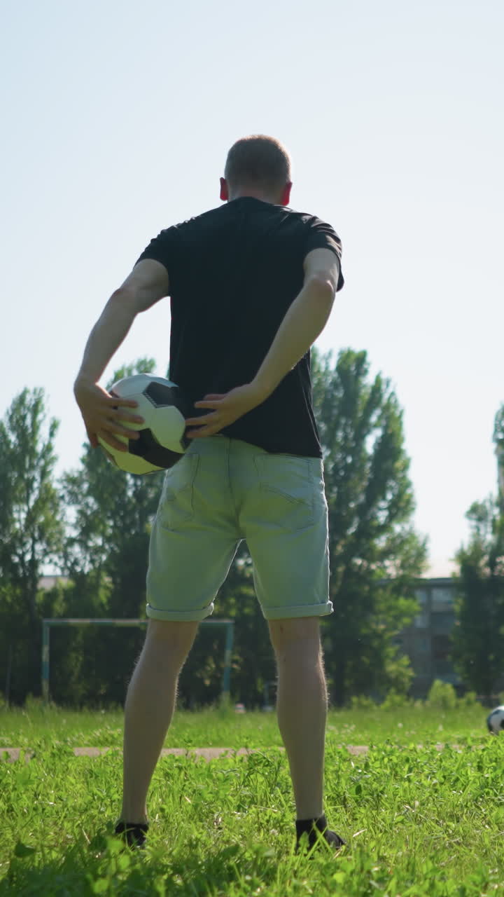 A close-up of a man rotating a soccer ball in his hands, with a view of people engaging in a soccer game in the background