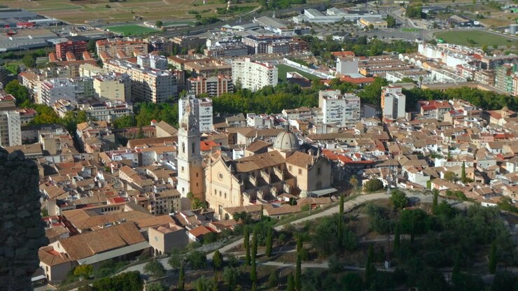 Veduta dall'alto di un denso quartiere cittadino spagnolo con torre di chiesa storica, cupola, tetti in tegole rosse, condomini, alberi verdi e terreno aperto in lontananza