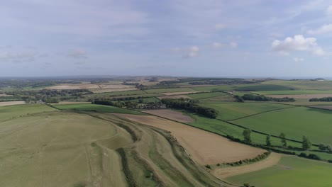 High-up-aerial-tracking-forward-over-Maiden-Castle-looking-south,-Sea-is-just-visible-on-the-horizon,-with-a-mixture-of-livestock-and-arable-fields-in-the-mid-ground