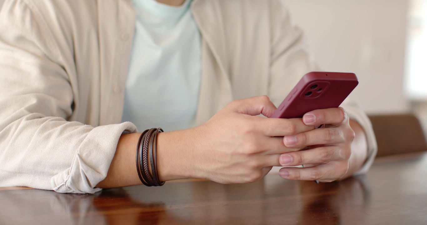 African American man 20s sitting at wood table adjusting red phone while scrolling with bracelets