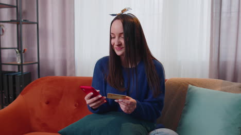 Woman-sitting-at-home-using-credit-bank-card-and-smartphone-while-transferring-money-online-shopping