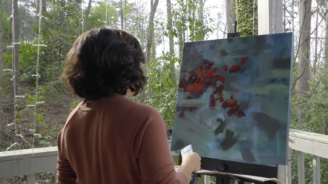 Female artist applies blue paint by moving brush across abstract multicolored painting on canvas and contemplates her work outside on a sunny day