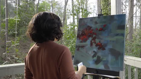 female artist applies blue paint by moving brush across abstract multicolored painting on canvas and contemplates her work outside on a sunny day
