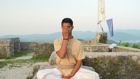 young indian male meditating at the top of the hill on ruins of castle wall doing yoga at sunrise in nature