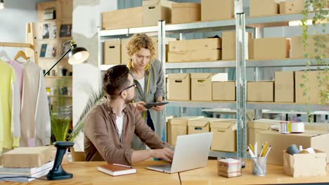 caucasian young professional designers working and speaking while browsing on laptop in clothing shop warehouse