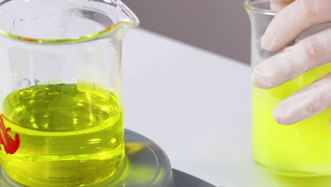 close-up of gloved hands manipulating beakers with bright yellow liquid on a lab table.