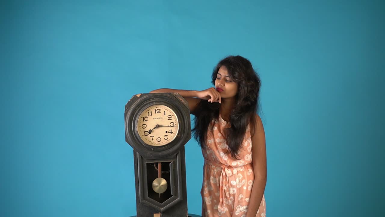 una joven india con vestido naranja parada con un viejo reloj vintage y pensando en un fondo azul aislado