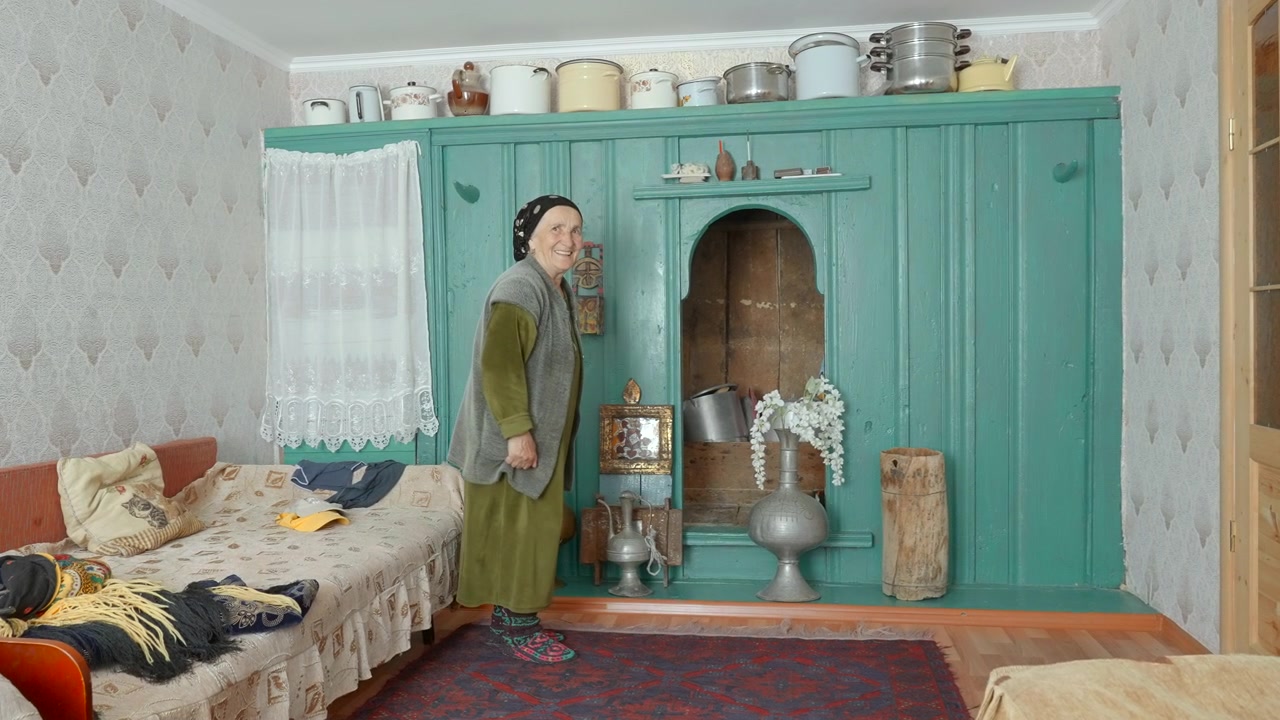Smiling woman stands beside turquoise cabinet in rustic home interior with decorative flowers, patterned wallpaper, lace curtain, stacked cookware, worn textures, and soft warm indoor light.