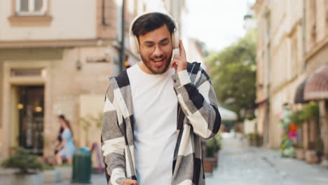 Hombre-árabe-Muy-Contento-Con-Auriculares-Inalámbricos-Escuchando-Música-Enérgica-En-Un-Teléfono-Inteligente-En-Una-Calle-De-La-Ciudad