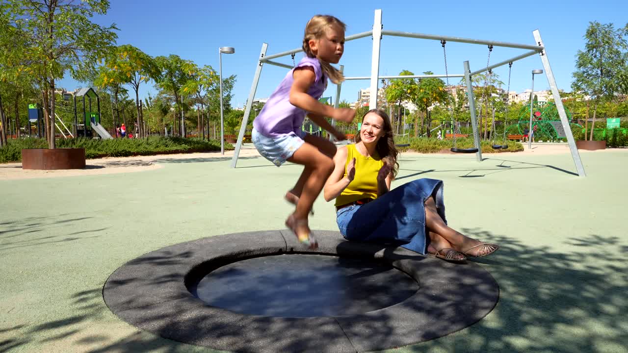 A girl jumping on a trampoline in a park with a woman watching