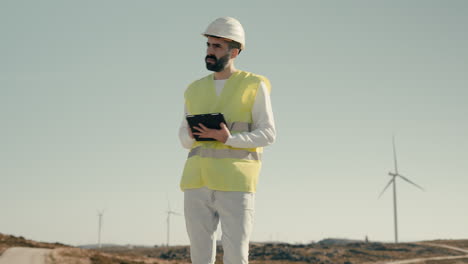 working to ensure a greener future, a male renewable energy engineer uses technology to audit wind turbines in a field of clean energy generators on a sunny day