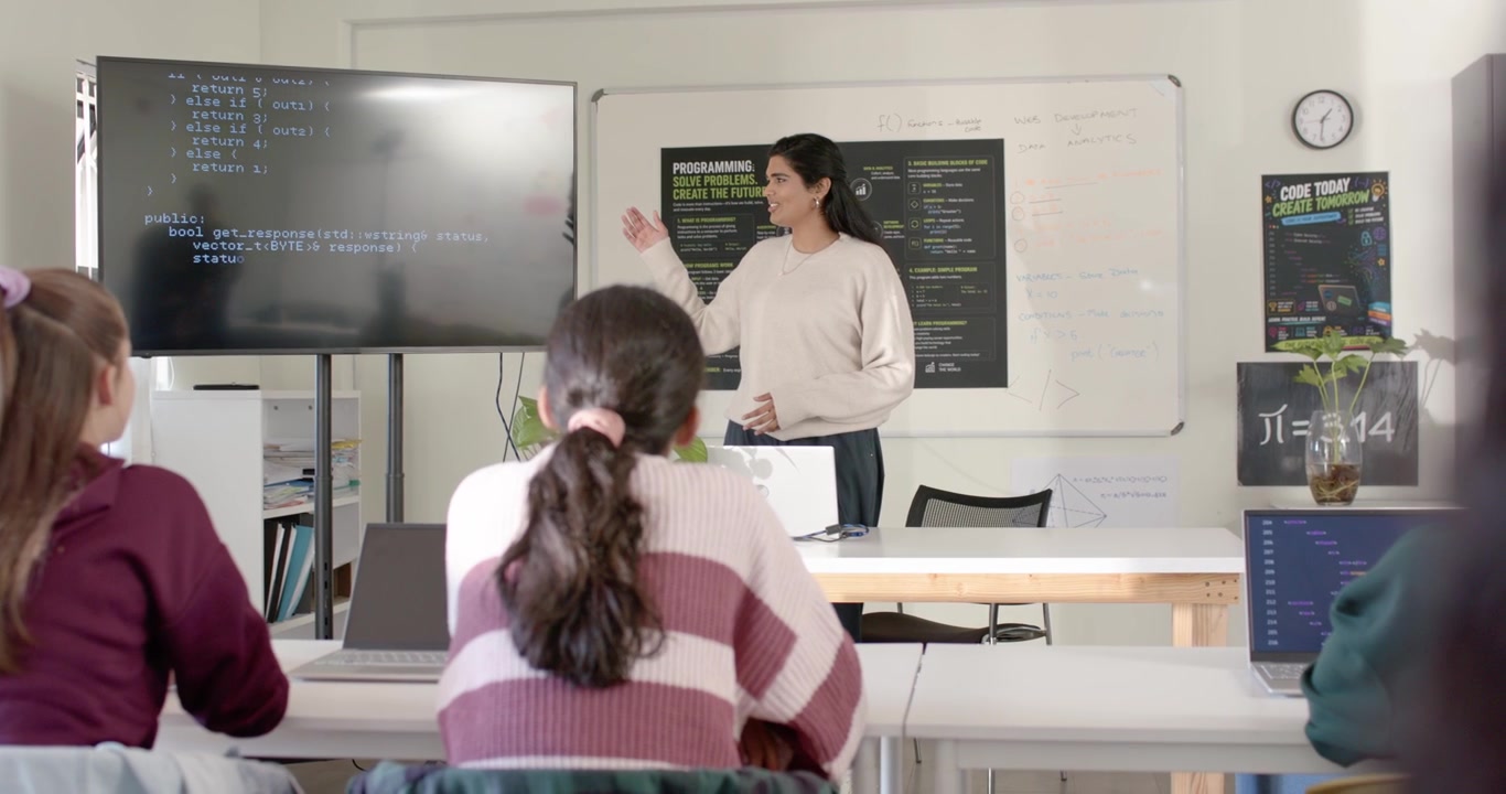 Indian teacher leading class, speaking and pointing to screen and laptops, teaching teens typing