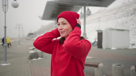 middle-aged woman in red hoodie adjusts her beanie outdoors during winter, warm smile on her face with blurred figures in background and urban structures visible under snowy conditions