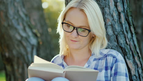 a blonde young woman in glasses reads a book in the park sits near a tree beautiful light before sun