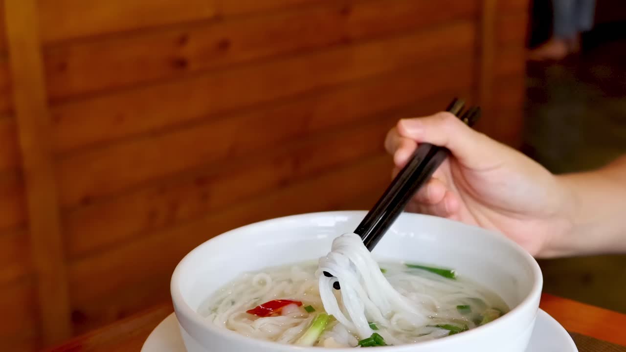 Close-up of hands using chopsticks to pick noodles and peppers from a bowl of soup.