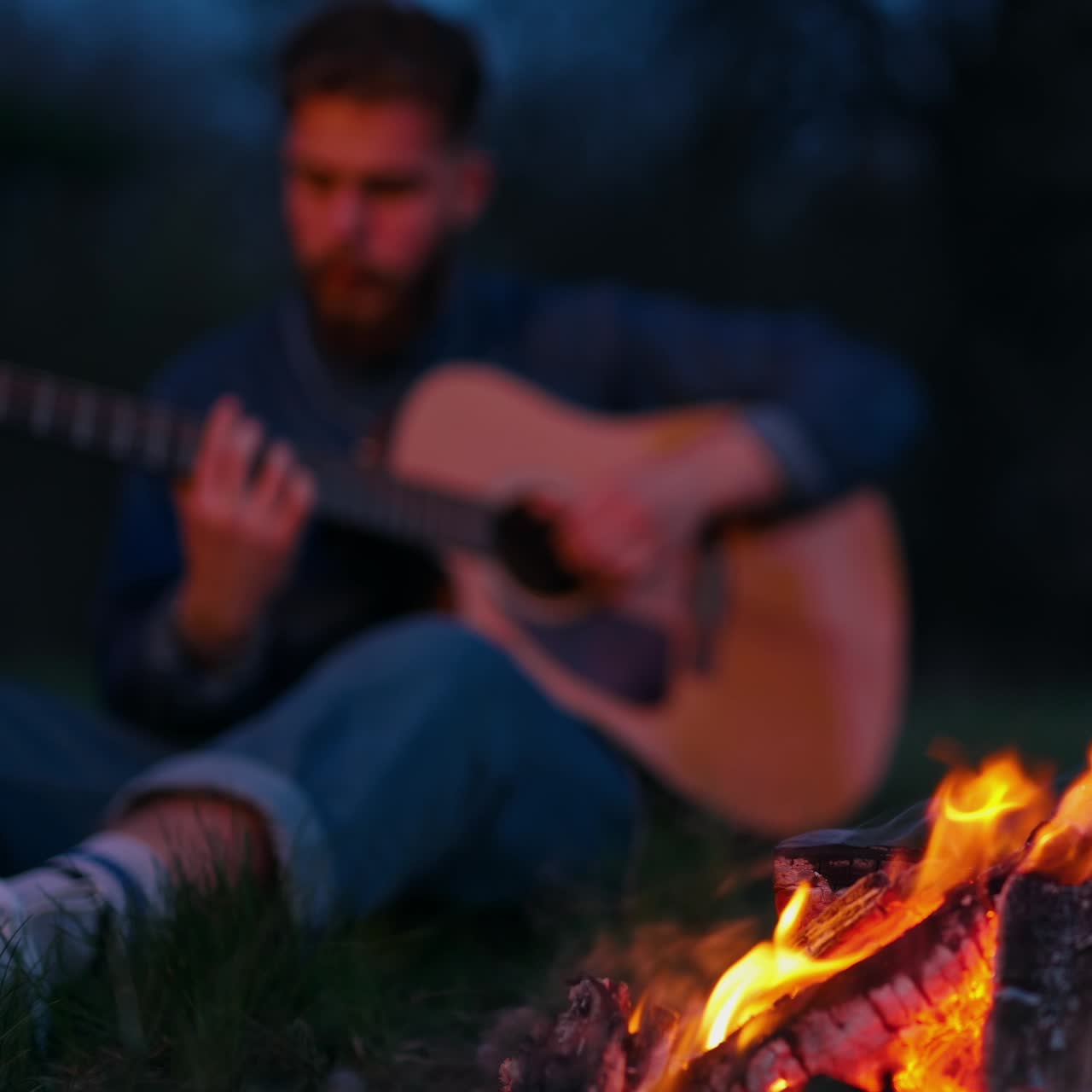 Man sitting by bonfire with guitar. Lonely man sitting by bonfire on the beach near forest playing acoustic guitar