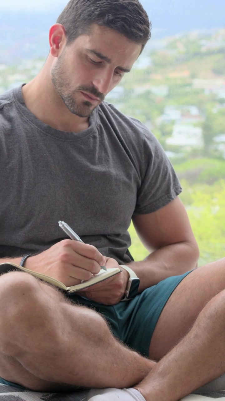 Video verticale: Uomo che controlla il respiro, con maglietta scura e pantaloncini verde acqua, scrive un diario in un quaderno sul balcone con una penna