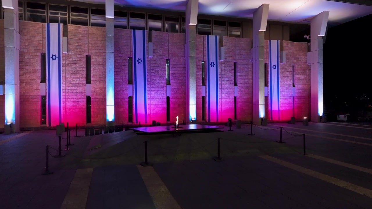 Facciata del parlamento Knesset illuminata con i colori nazionali, con fiamma commemorativa eterna e bandiere appese verticalmente a Gerusalemme di notte.