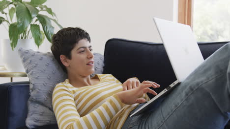 Happy-caucasian-woman-lying-on-sofa,-using-laptop-and-smiling-in-sunny-house