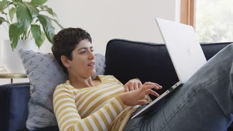 feliz mujer caucásica acostada en el sofá, usando una computadora portátil y sonriendo en una casa soleada