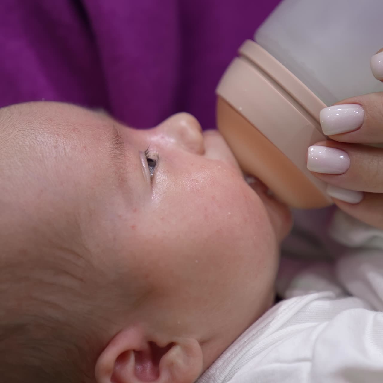 Infant babe is being fed by his mum from a bottle. Kid suckling milk from the bottle and slowly falling asleep. Close up