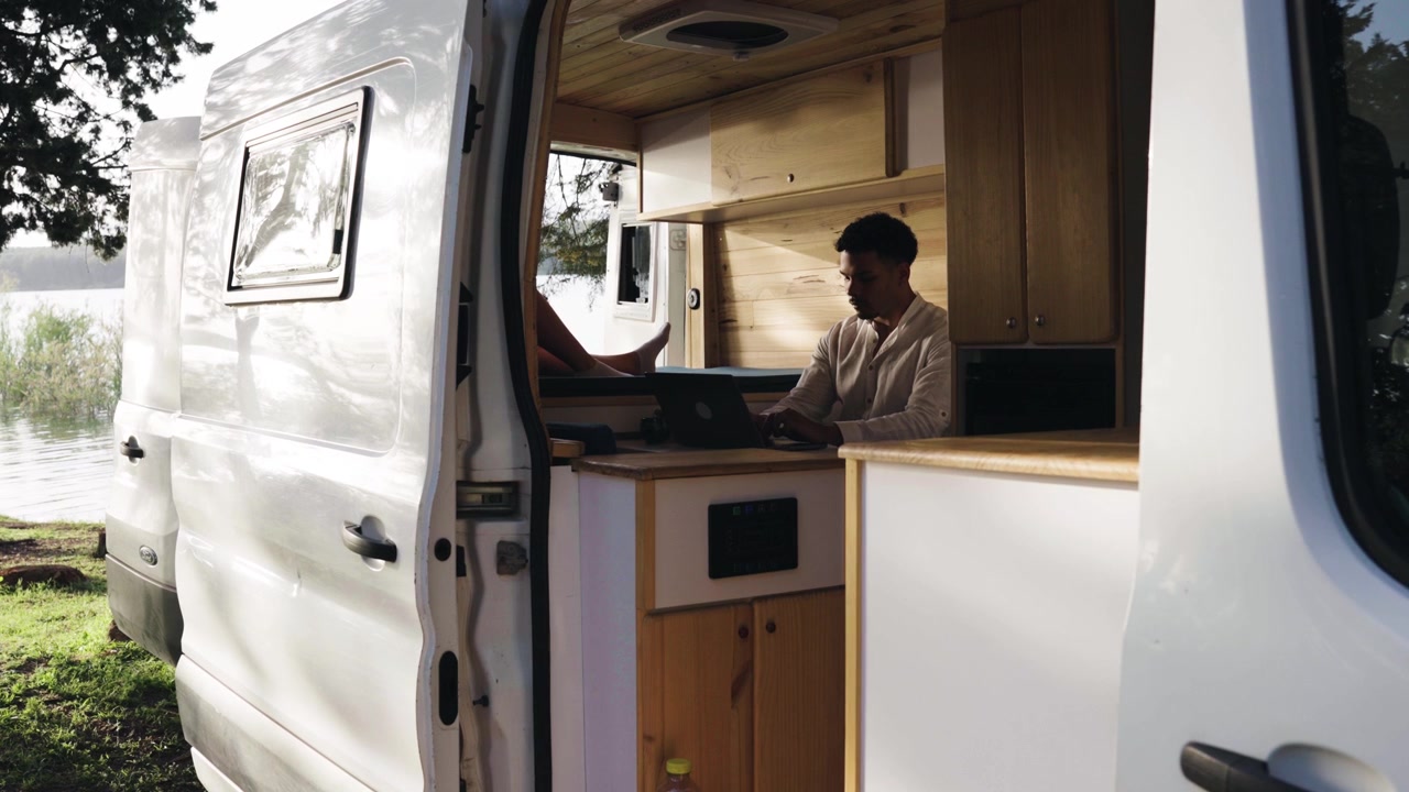 Giovane uomo che lavora al laptop all'interno di un camper in riva a un lago