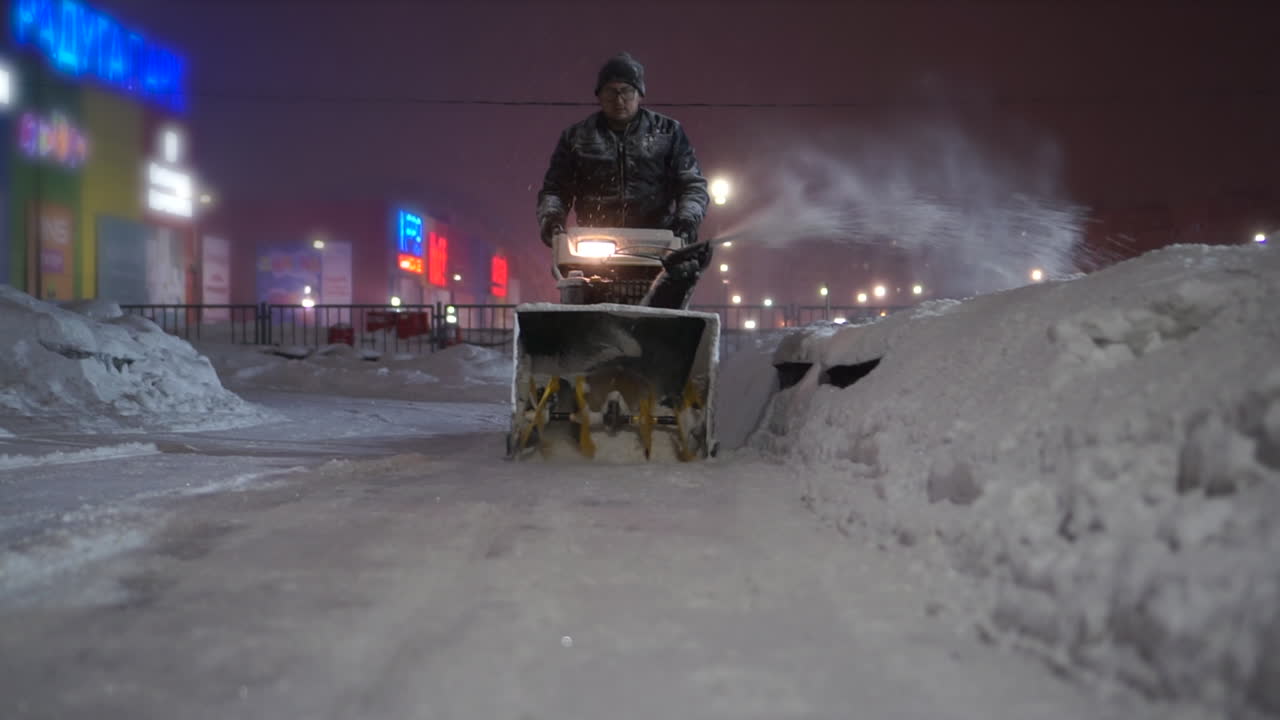 Snowblower clearing snow at night near a shopping mall