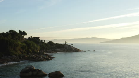 Vista-Aérea-De-Drones-Sobre-El-Agua,-Olas-Del-Mar-Y-Puesta-De-Sol-En-El-Océano,-Una-Fantástica-Costa-Rocosa-Con-Niebla-Y-Montañas-De-Selva-Tropical-En-El-Horizonte,-Jurere-Internacional,-Florianópolis,-Santa-Catarina,-Brasil