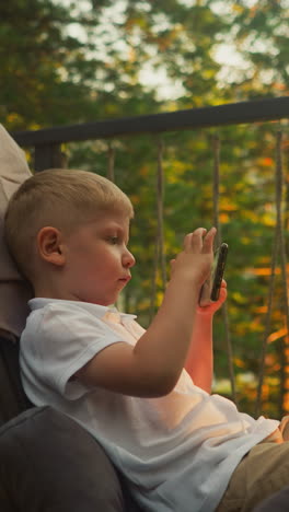 boy plays game on mobile phone sitting in armchair on balcony with incredible view of nature beauty. luxurious glamping in country house slow motion