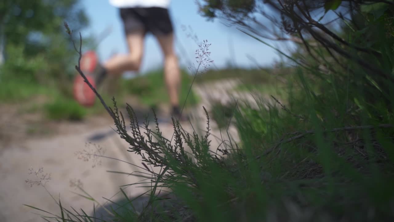 A shot of an out of focus recreational trail runner jogging along a remote gravel road on a summer's day. Foreground capturing a close up of the grass and leaves of the remote landscape.